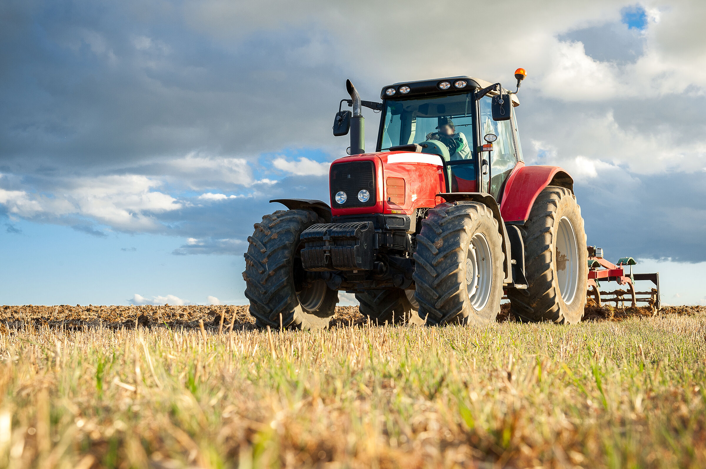 Wie kommen die billigen Preise bei Lidl & Co.zustande? Zahlen die Bauern drauf? (Foto: David San Segundo/adobe.stock.com) Disounter-Bio: Ein roter Traktor fährt auf dem Feld, über ihm wolkiger Himmel